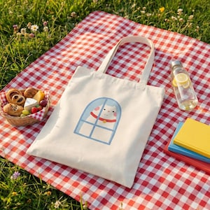 以下が含まれることがあります： 青い窓枠の中に猫のイラストが描かれた白いキャンバストートバッグ。赤と白のチェック柄のピクニックブランケットの上に置かれています。食べ物のバスケット、ウォーターボトル、本もブランケットの上にあります。