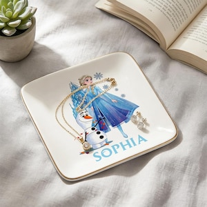 Może przedstawiać: Biała kwadratowa patera na drobiazgi ze złotą obwódką, z grafiką Elsy i Olafa z Krainy Lodu. Złoty naszyjnik i kolczyki spoczywają na paterze, z imieniem "SOPHIA" w niebieskim tekście.