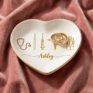 May include: Heart-shaped white ceramic ring dish with gold trim, featuring gold medical symbols and two gold rings. The name "Ashley" is scripted in gold. The dish rests on a pink velvet surface.