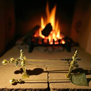Pode incluir: Um conjunto de ferramentas para lareira em latão dourado, incluindo uma pá, um atiçador e um suporte. As ferramentas têm detalhes decorativos. O fundo mostra uma lareira com fogo a arder.