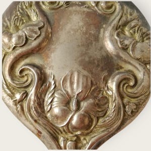 May include: Close-up of a silver-toned, ornate compact mirror. The design features floral and scrollwork motifs, with a central flower and surrounding embellishments. The metal has a slightly aged appearance.