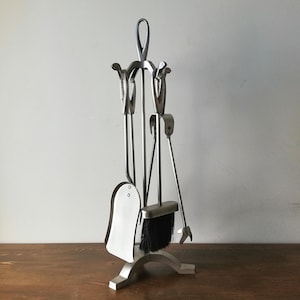 Puede incluir: Un juego de herramientas para chimenea plateado con soporte. El juego incluye una pala, un cepillo, un atizador y unas pinzas. El cepillo tiene cerdas negras. Las herramientas son de metal y tienen un diseño elegante y moderno. El soporte es curvo en la base.