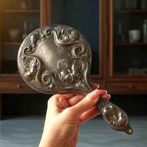 May include: An antique silver-toned hand mirror with floral relief detailing. The oval mirror features ornate floral designs around the edges and on the handle. The mirror is held by a hand, showcasing the intricate craftsmanship of the vintage piece.