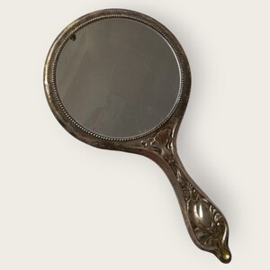 May include: An antique hand mirror with a round reflective surface and a decorative handle. The mirror's frame and handle are silver-toned, with ornate floral detailing on the handle. The mirror is approximately 5 inches in diameter.