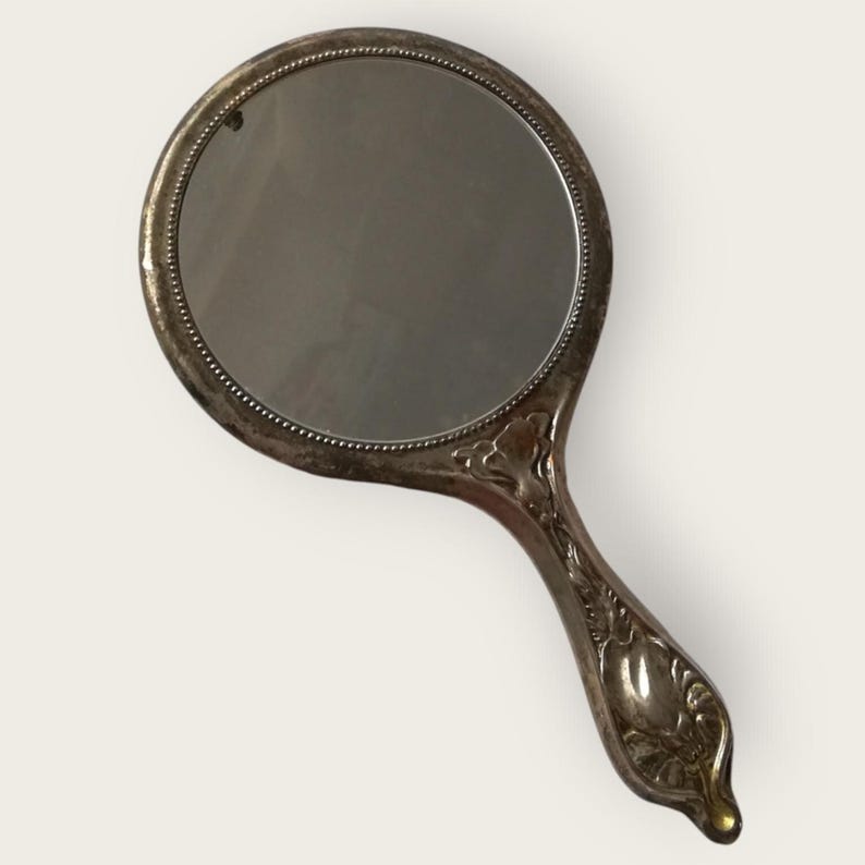 May include: An antique hand mirror with a round reflective surface and a decorative handle. The mirror's frame and handle are silver-toned, with ornate floral detailing on the handle. The mirror is approximately 5 inches in diameter.
