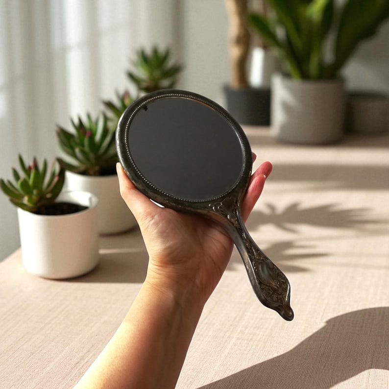 May include: An antique hand mirror with an oval-shaped reflective surface and an ornate, decorative handle. The mirror's frame and handle have a dark, metallic finish. The handle features floral and scrollwork designs. The mirror is held in front of a neutral background.