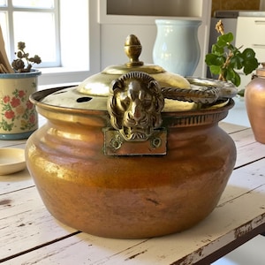 Puede incluir: Una olla de cobre con tapa de latón y asa con cabeza de león. La olla tiene forma redonda y un acabado deslustrado. La tapa está rematada con un adorno decorativo. La olla está sobre una mesa de madera blanca.