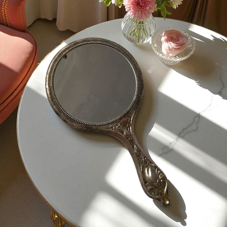 May include: An antique hand mirror with a silver-colored frame and handle. The round mirror has a beaded edge and is set against a white marble-like surface. The handle features an ornate floral design.