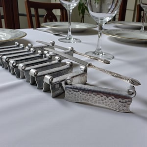 May include: A set of silver-colored knife rests with a hammered texture, arranged on a white tablecloth. Two knives rest on the holders, near plates and wine glasses, suggesting a formal dining setting.