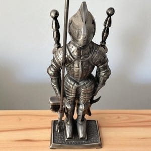 Könnte beinhalten: Ein silberfarbenes Metall-Kaminbesteck mit einem Ritter in voller Rüstung. Das Set enthält einen Schürhaken, eine Bürste und andere Werkzeuge, die alle auf einem rechteckigen Sockel montiert sind. Die Ritterfigur ist detailliert.