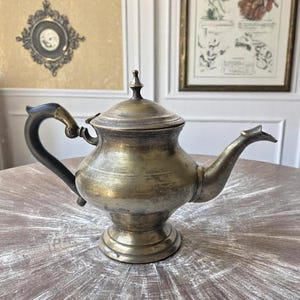Pode incluir: Bule de latão antigo com uma alça escura e um bico decorativo. O bule tem uma tampa com um pequeno pináculo e assenta sobre uma base redonda. O bule é exibido em uma mesa de madeira.