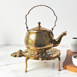 Könnte beinhalten: Eine Vintage-Teekanne aus Messing mit floralem Muster und gebogenem Griff. Die Teekanne steht auf einem passenden Messing-Trivet mit Zierausschnitten und drei Beinen. Die Teekanne und das Trivet sind goldfarben.