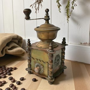 Puede incluir: Antiguo molinillo de café de latón y madera con manivela. El molinillo tiene una base cuadrada con detalles florales decorativos y un pequeño cajón. Granos de café esparcidos sobre una superficie de madera alrededor del molinillo.