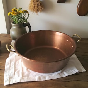 May include: A large, round copper bowl with brass handles rests on a white cloth. The bowl has a polished finish. A small vase with yellow flowers is in the background, along with other kitchen items.