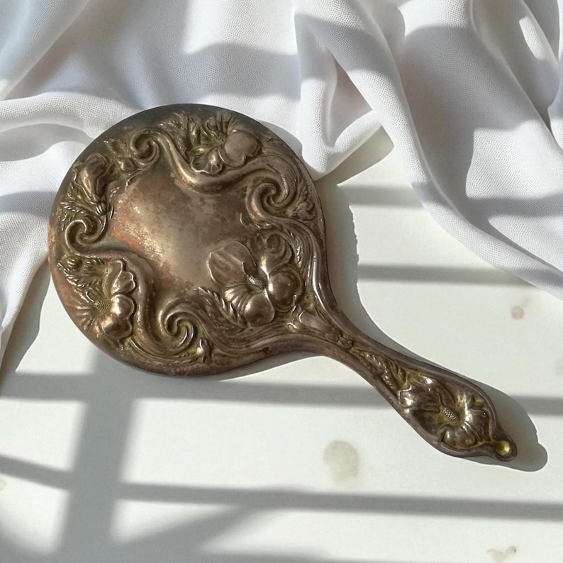 May include: An antique silver-toned hand mirror with an ornate floral design. The oval mirror has a decorative handle and is resting on a white surface. The mirror's surface shows signs of age and use.
