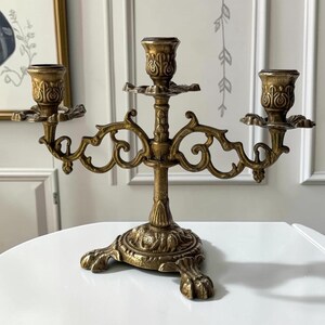 May include: A three-armed brass candelabra featuring ornate scrollwork arms, three candle cups with embossed detailing, and a sturdy base supported by three claw-style feet. The metal surface has a textured, aged gold-toned finish throughout.