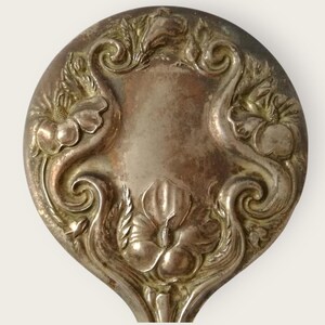 May include: Close-up of a silver spoon head with an ornate floral design. The spoon features a central shield shape surrounded by swirling patterns and detailed flower motifs. The metal has a slightly tarnished appearance.