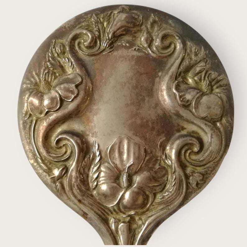May include: Close-up of a silver spoon head with an ornate floral design. The spoon features a central shield shape surrounded by swirling patterns and detailed flower motifs. The metal has a slightly tarnished appearance.