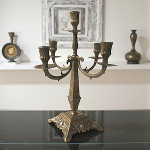 Può includere: Un candelabro vintage in ottone con cinque portacandele. Il candelabro ha una base ornata e bracci decorativi. Il design generale è simmetrico e l'ottone ha una calda patina invecchiata. Un elemento decorativo per la casa.