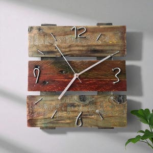 May include: Square wooden wall clock with a rustic design. The clock face is made of three horizontal wooden planks in shades of brown and red. Silver metal numerals and hands indicate the time. The clock is mounted on a light grey wall.