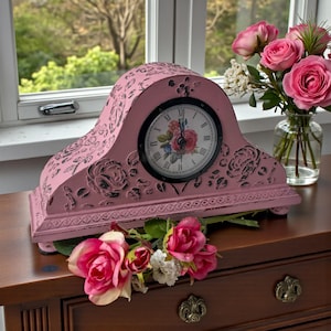 May include: A pink mantel clock with a floral design and Roman numerals. The clock has a distressed finish and is decorated with rose motifs. A bouquet of pink roses sits nearby.