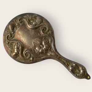 May include: An antique silver-toned hand mirror with floral and scrollwork detailing. The oval mirror features embossed flowers and swirling patterns on the back and handle. The handle has a rounded end.