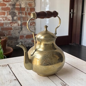 May include: A vintage brass teapot with a dark wooden handle and a curved spout. The teapot has an etched floral design on its body and a small lid with a knob. The handle is attached to the body with brass supports. The teapot is sitting on a wooden surface.