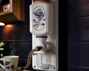May include: A wall-mounted coffee grinder featuring a white ceramic hopper with a floral rose decal, a wooden lid, a metal hand crank with a wooden knob, and a clear glass catch container mounted on a white rectangular metal backplate.