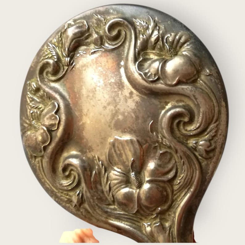 May include: Antique silver-toned hand mirror with an oval shape. The mirror's surface is surrounded by an embossed floral design featuring hibiscus flowers and swirling vines. The handle is not visible in the image.