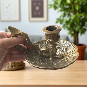 Könnte beinhalten: Ein Vintage-Kerzenhalter aus Messing mit aufwendigen Reliefdetails. Der Halter hat eine zentrale Tasse für die Kerze, einen dekorativen Sockel mit Figuren und einen Griff mit stilisiertem Design. Die Farbe ist warmes, gealtertes Messing.
