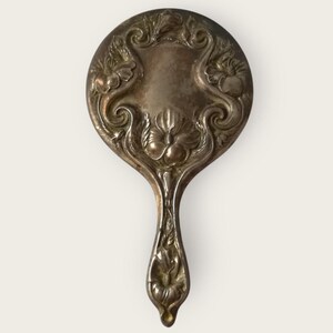 May include: Antique silver-toned hand mirror with an ornate floral design. The round mirror features detailed relief carvings of flowers and swirling patterns around the edges and on the handle. A vintage vanity accessory.