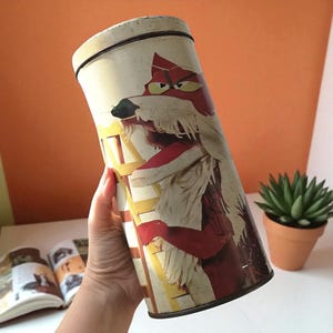 May include: Vintage cylindrical tin featuring a cartoon fox illustration in red, white, and brown. The tin has a white background with yellow and brown accents. The tin is held by a person's hand.