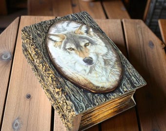 May include: A rectangular wooden box with a hinged lid. The lid features an oval portrait of a wolf with gray and brown fur. The box has a textured, bark-like appearance with gold accents. The box is sitting on a wooden surface.