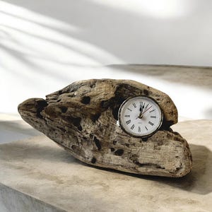 May include: A small clock with a white face and black Roman numerals is set into a piece of weathered driftwood. The clock has a gold-coloured bezel and red second hand. The driftwood has a natural, textured appearance.