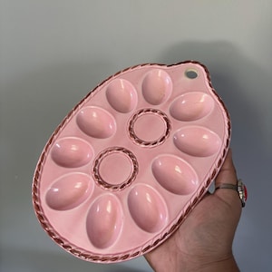 Puede incluir: Una bandeja de cerámica para servir huevos de color rosa claro con forma ovalada, que presenta diez hendiduras con forma de huevo dispuestas alrededor de una sección circular central. Tiene un borde texturizado de cuerda en tono rosa oscuro.
