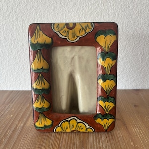 以下が含まれることがあります： 赤色の縁取りに、黄色と緑の花柄が施された長方形のセラミックフォトフレーム。 フレームの内側はクリーム色で、木製の表面に展示されています。 フレームの高さは約15cm、幅は約10cmです。