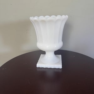 May include: A white milk glass vase featuring a scalloped rim, vertical fluted detailing along the body, and a pedestal base that transitions into a square-shaped foot. The vessel has a smooth, opaque finish and a wide, flared opening.