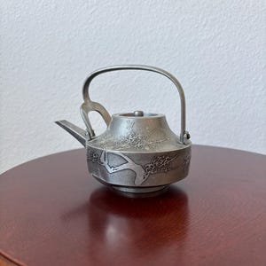 May include: A small, silver-colored teapot with a curved handle and a spout. The teapot is decorated with an embossed floral design. It sits on a round, dark brown wooden table.