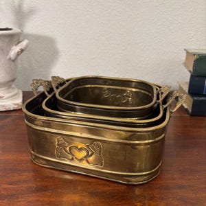 May include: A set of three brass planters of different sizes. The largest has a heart and hands design. Each planter has decorative handles. The brass has a warm, golden hue, and the planters are nested together.