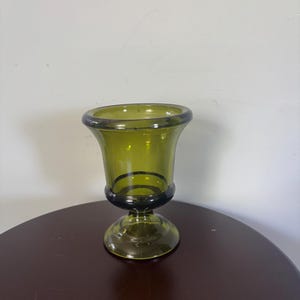 May include: A translucent olive green glass vase featuring a wide, flared rim and a rounded bowl shape that tapers down to a thick, circular pedestal base. The glass has a smooth, polished finish with a consistent deep green hue throughout the structure.