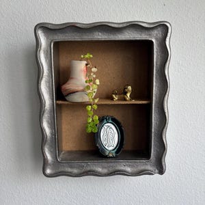 May include: A rectangular wall shelf with a wavy, metallic silver frame and two interior shelves. The top shelf holds a small, marbled pink and white vase with a trailing green plant and a brass dog figurine. The bottom shelf contains a small, oval, dark-framed mirror with a decorative etched design.