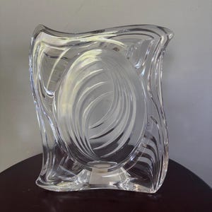 Peut inclure: Un cadre photo en verre transparent avec un bord extérieur ondulé et asymétrique et une ouverture ovale centrale. Le cadre présente des lignes courbes gravées rayonnant depuis le centre, créant un motif tourbillonnant dans le verre épais.