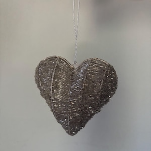 May include: A heart-shaped hanging ornament covered in metallic silver beads. The surface features a textured, ribbed pattern created by the dense arrangement of small, reflective beads. A thin silver cord is attached to the top for hanging.