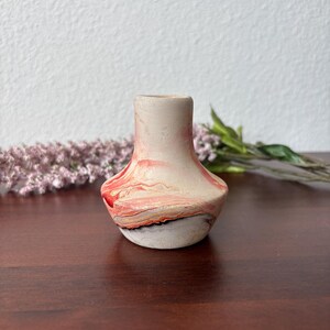 May include: A small, decorative vase with a unique, marbled design. The vase features a light pink base with swirls of red, gray, and black. The vase has a wide base and a narrow neck, perfect for displaying small flowers or as a decorative accent.