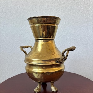 Puede incluir: Una cafetera de latón vintage con textura martillada. La cafetera tiene una base bulbosa, dos asas y una parte superior acampanada. Se apoya sobre tres patas de garra. El latón tiene un tono dorado cálido, lo que sugiere antigüedad y uso. El diseño recuerda a los utensilios de cocina antiguos.