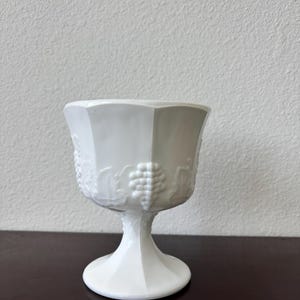 Puede incluir: Un cáliz blanco de estilo vintage con base de pedestal. El vidrio presenta un diseño en relieve de uvas y hojas alrededor del cuenco. El cáliz tiene una forma geométrica facetada y probablemente está hecho de vidrio o cerámica.