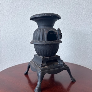 May include: A small, black cast iron potbelly stove replica featuring a rounded, ribbed body, a circular top plate with decorative edge detailing, a small hinged door on the front, and four curved legs extending from a square base.