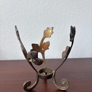 May include: A vintage metal egg holder with a floral design. The holder has a circular base supported by three curved legs. The top features stylized leaves and a central support for an egg. The metal has a tarnished, antique appearance.