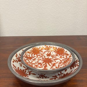 Puede incluir: Juego de dos platos de cerámica. El juego incluye un cuenco pequeño dentro de un plato más grande. Ambas piezas presentan un fondo blanco con un estampado floral rojo y un borde gris. El cuenco pequeño tiene detalles dorados.