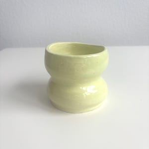 May include: A small, pale yellow ceramic cup with a smooth, glossy finish. The vessel features a distinct hourglass shape with a wide, rounded base, a narrow middle section, and a slightly flared, uneven rim at the top opening.
